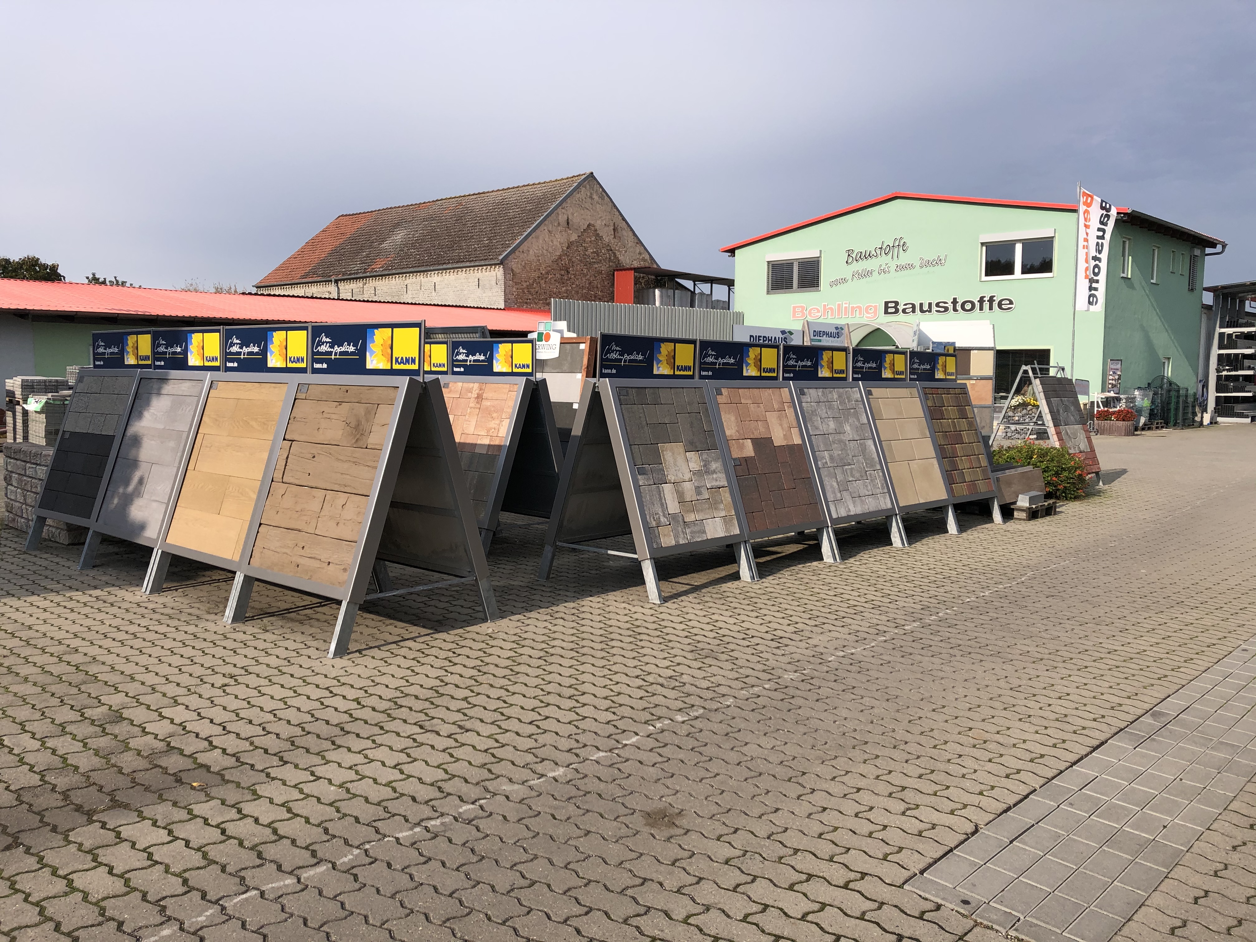 Reihen von verschiedenen Bodenfliesen aufgeständert im Freigelände eines Baumarkts, umgeben von einer grünen Gebäude und Lagerschildern
