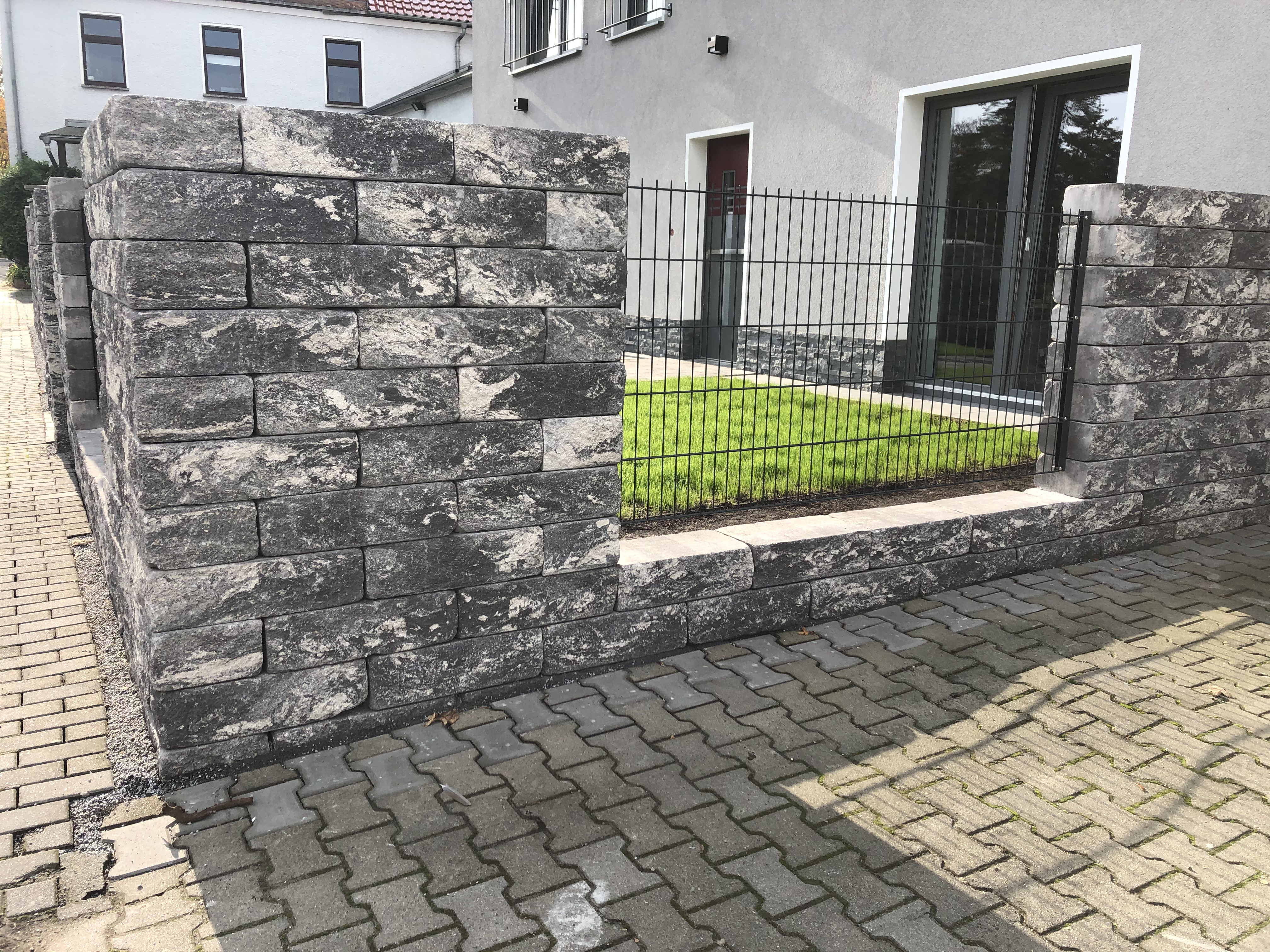 Gartenmauer aus grauem Naturstein mit integriertem Metallzaun vor einem modernen Gebäude mit gepflastertem Gehweg