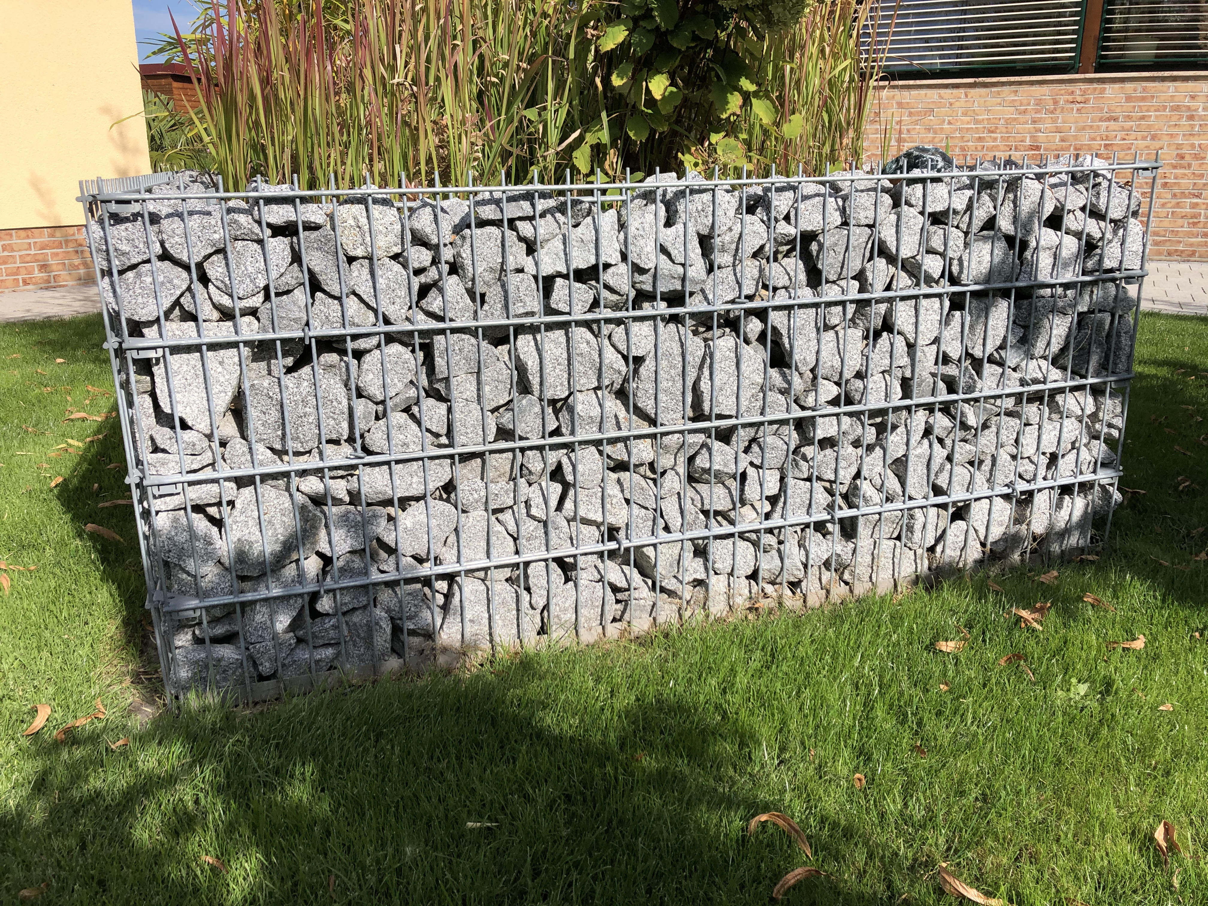 Eine Gabionenwand aus Steinen in einem sonnigen Garten, umgeben von Gras und Pflanzen.