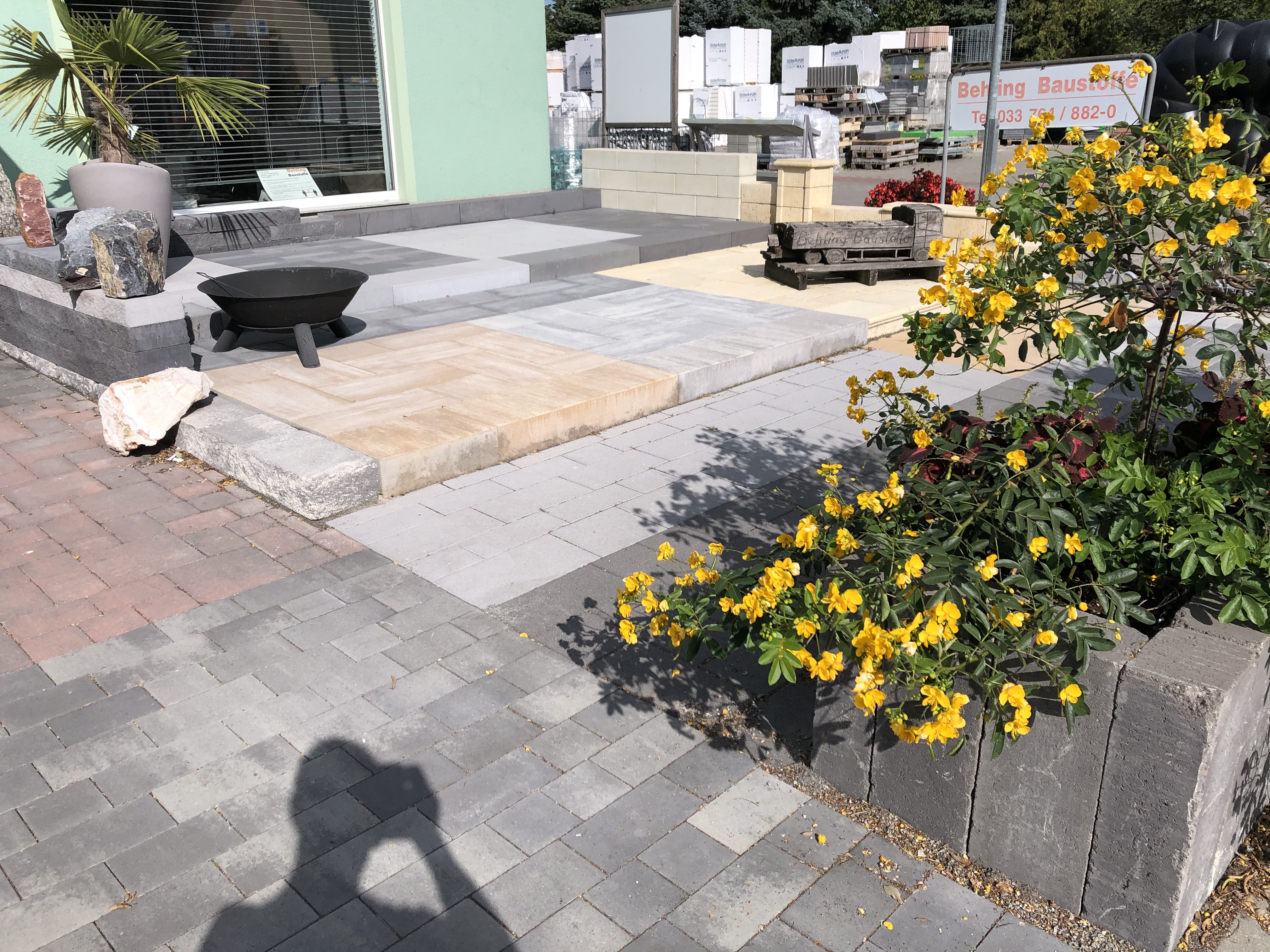 Terrassengestaltung mit grauen und beigefarbenen Pflastersteinen, Feuerschale, gelben Blumen und Palmen in einem Außenbereich