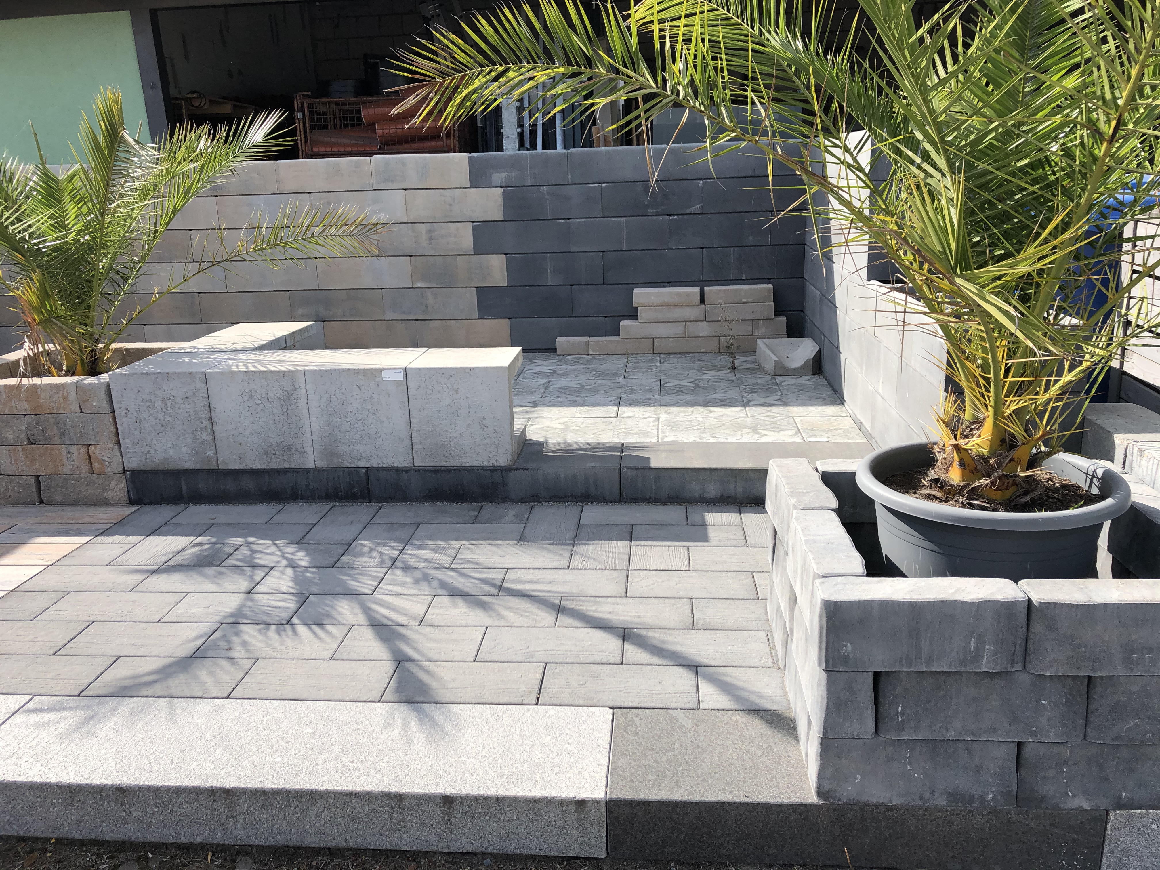 Eine moderne Terrasse mit grauen Pflastersteinen, erhöhten Betonblöcken und Palmen in Pflanzkübeln.