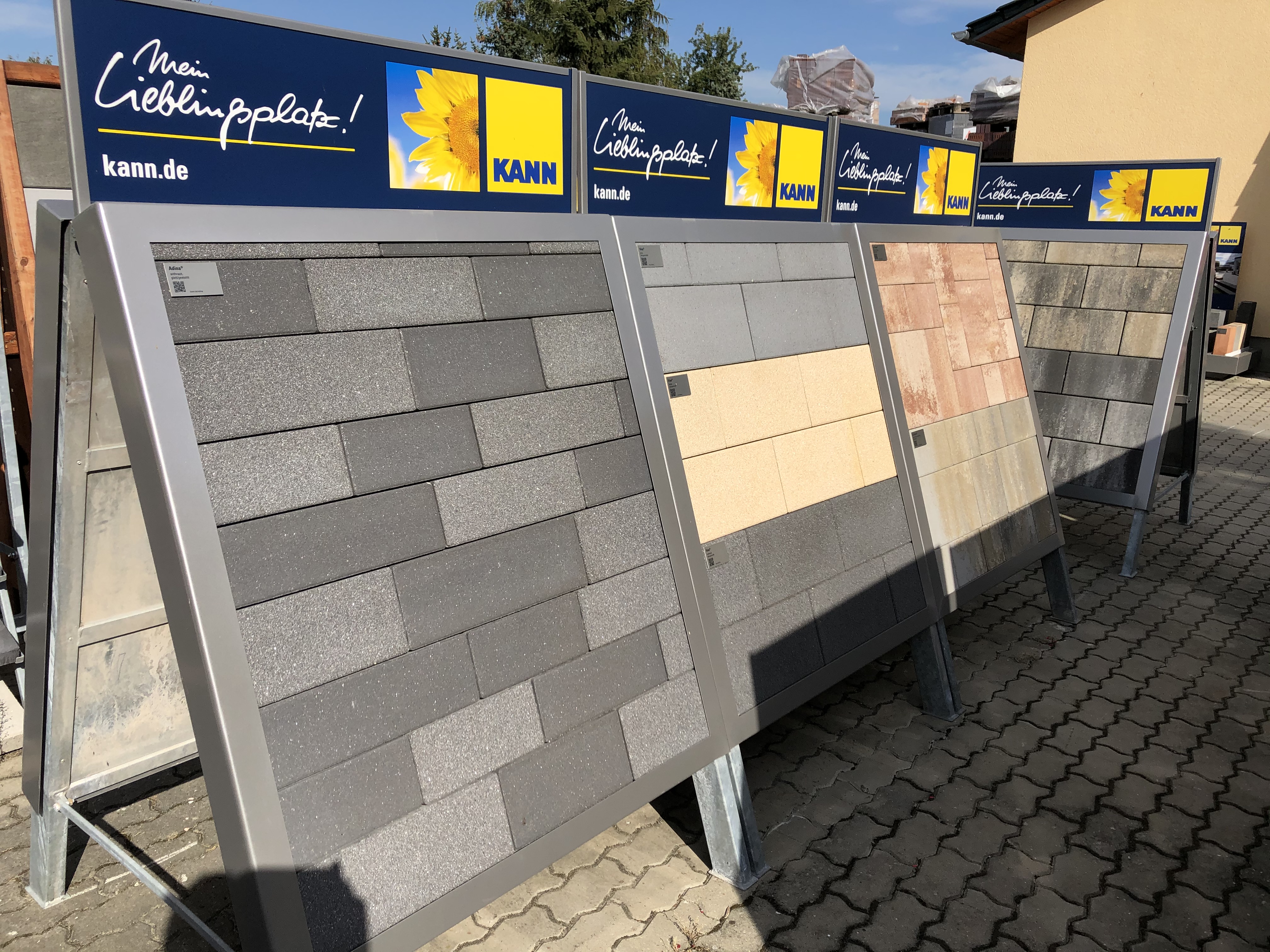 Verschiedene Terrassenplatten auf Metallständern zur Ansicht im Außenbereich ausgestellt. Verschiedene Farben und Oberflächentexturen zu sehen.