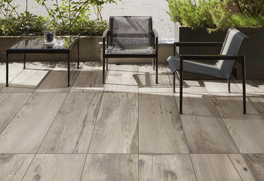 Stilvoll eingerichtete Terrasse mit zwei schwarzen Sesseln, einem Tisch mit Glasplatte und großen hellen Fliesen, umgeben von grünen Pflanzen.