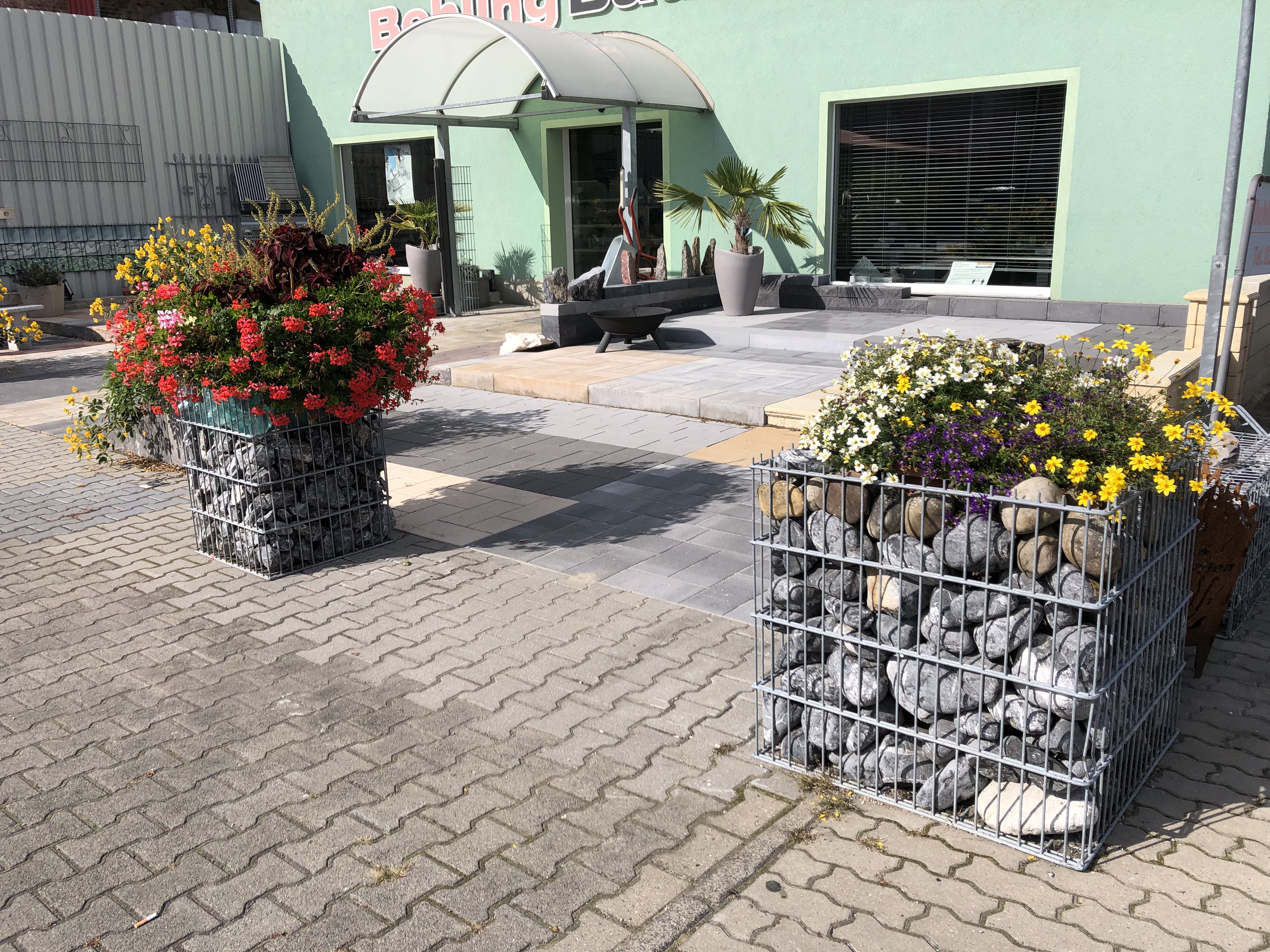 Eingangsbereich eines Gebäudes mit Gabionen, die mit bunten Blumen bepflanzt sind.