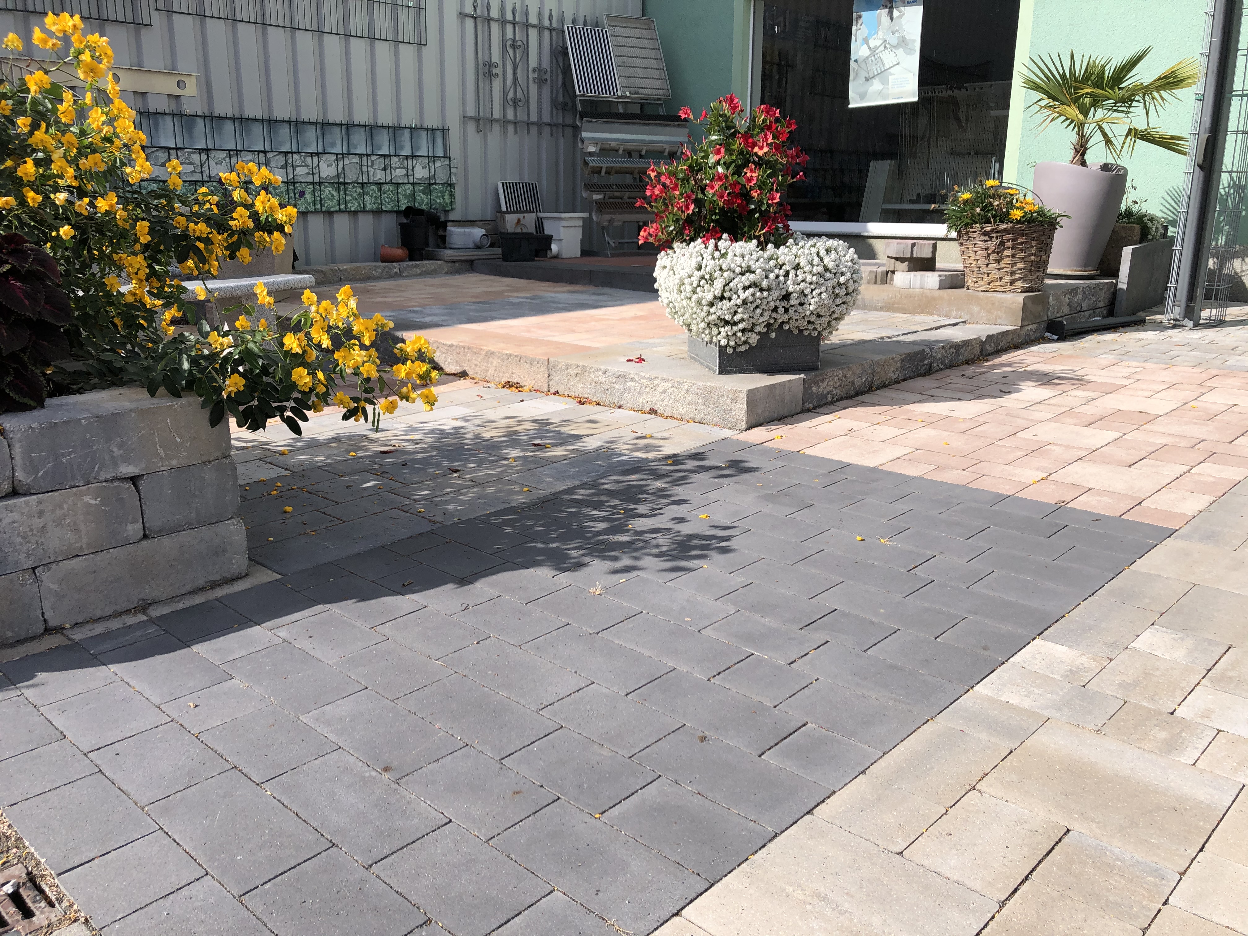 Gepflasterter Außenbereich mit bunten Blumen und Pflanzen in Töpfen, darunter gelbe und rote Blüten, sowie ein grauer und ein beige gepflasterter Weg.